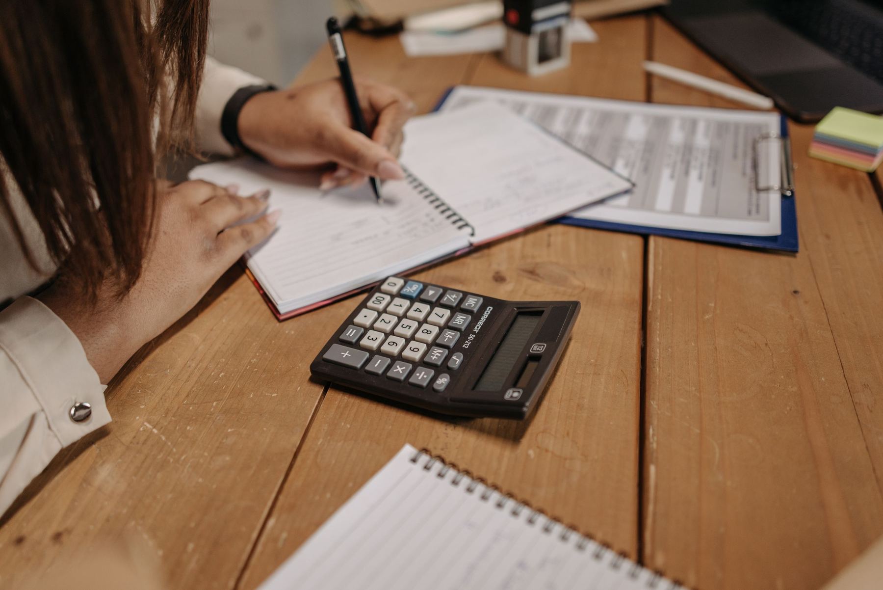 Hands writing figures in a notebook beside a calculator and paperwork